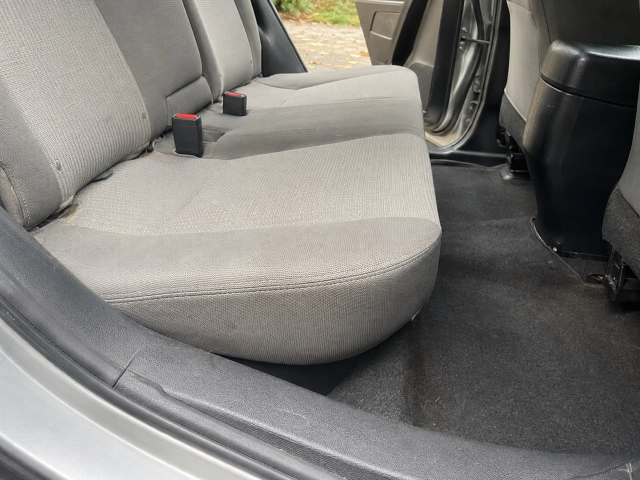 A bare grey cloth rear bench seat photographed from the open door: the cushion slopes back toward the seatback, a seam runs along where they meet, and past the front edge the carpeted footwell drops away.